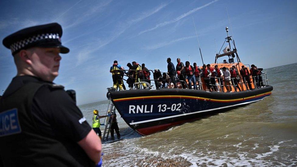 A British police officer stands guard on the beach of Dungeness, on the southeast coast of England, on June 15, 2022, as Royal National Lifeboat Institution's (RNLI) members of staff help migrants to disembark from one of their lifeboat after they were picked up at sea while attempting to cross the English Channel.