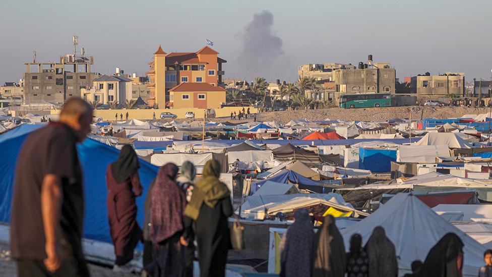 Smoke rises in the distance while people stand in front of a tent community in Rafah on 7 May