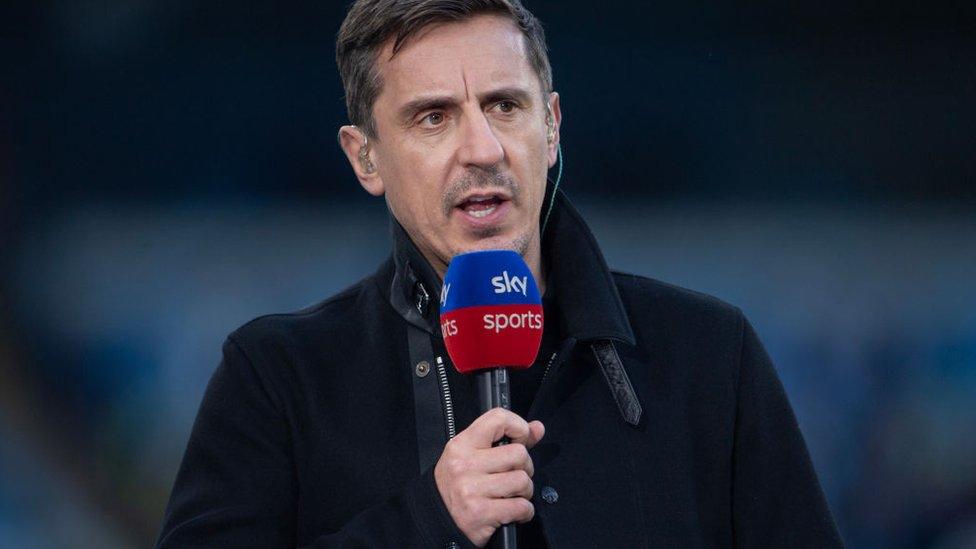 Gary Nevill stands in a football stadium wearing a thick winter coat and holding a microphone, which has a blue and red windshield with Sky Sports' logo printed on it in white. He is also wearing an ear piece made of translucent plastic in each ear, and a clear plastic wire can be seen trailing down into the collar of his coat. He is looking off to the side and appears to be speaking. He's got short black hair and faint stubble on his top lip and chin.