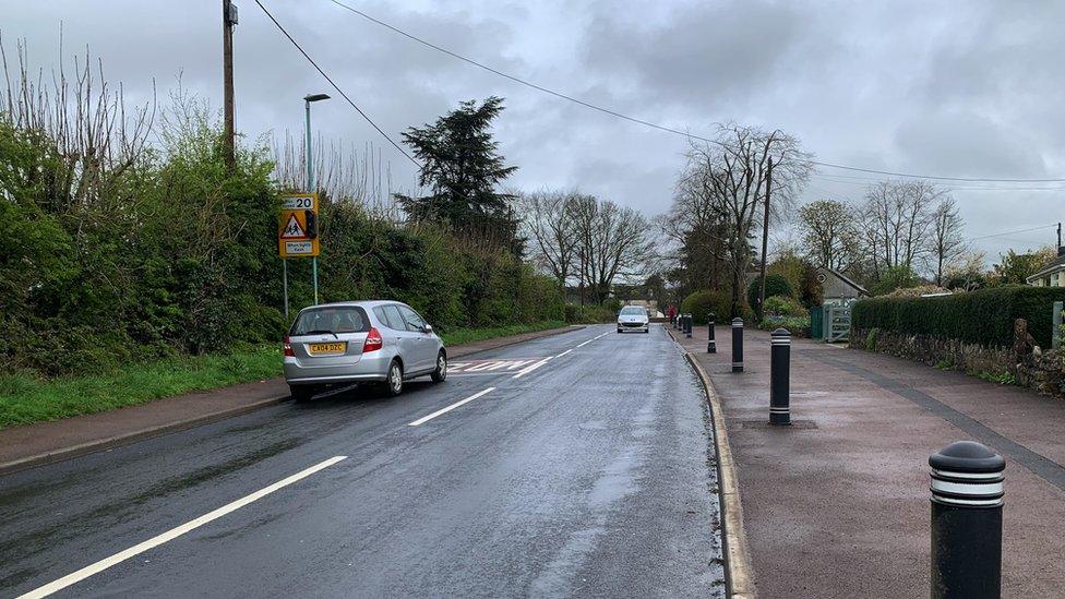 Image of the road by Tutshill C of E Primary School. A school sign can be seen, as well as bollards on the pavement.