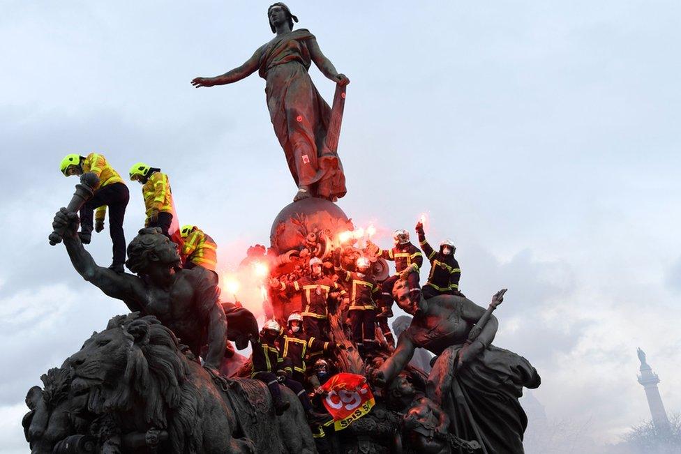 Firefighters brandish flares as they climb on the Statue of Republic Triumph at Nation square in Paris during a demonstration to protest against French government's plan to overhaul the country's retirement system