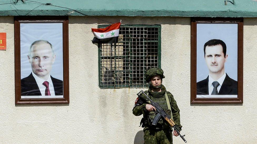 A member of the Russian military stands outside a building in Syria between portraits of Vladimir Putin and Bashar al-Assad.