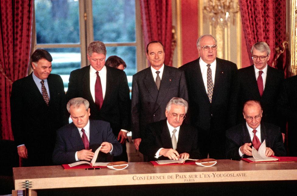 Presidents sit at a table and sign a peace agreement as world leaders watch from behind