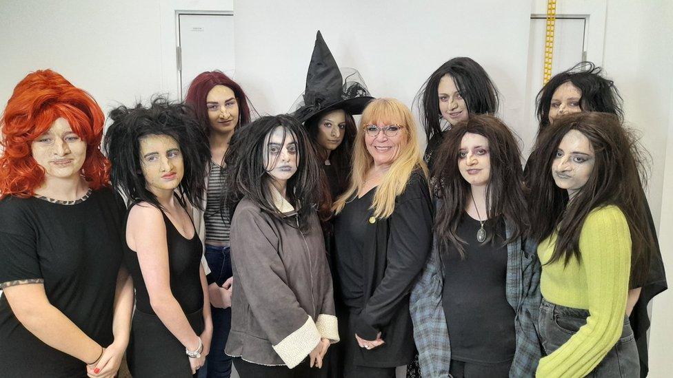 Woman with long blonde hair with group of students with long dark hair and make-up. One is wearing a witch's hat.