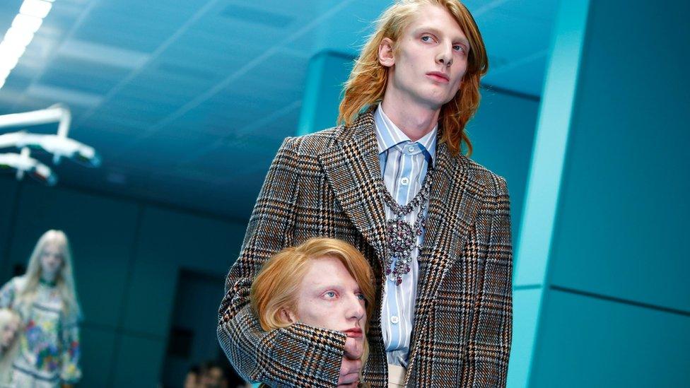 Male model walks down the catwalk holding a mannequin head that is a replica of his head