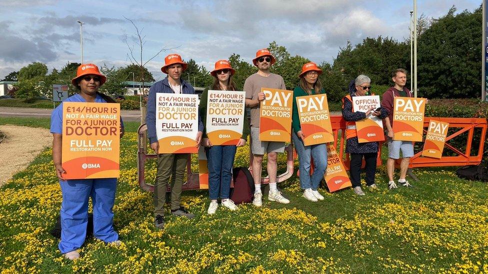 Seven people standing outside holding signs about fair pay for doctors, all with orange bucket hats on