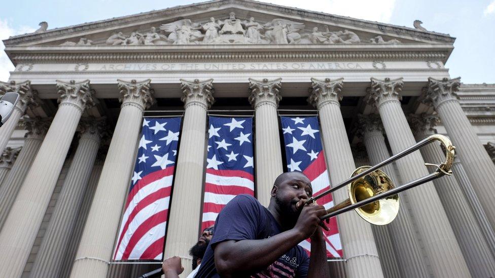 A man is pictured playing the trombone in front of US Archives building, adorned in hanging flags