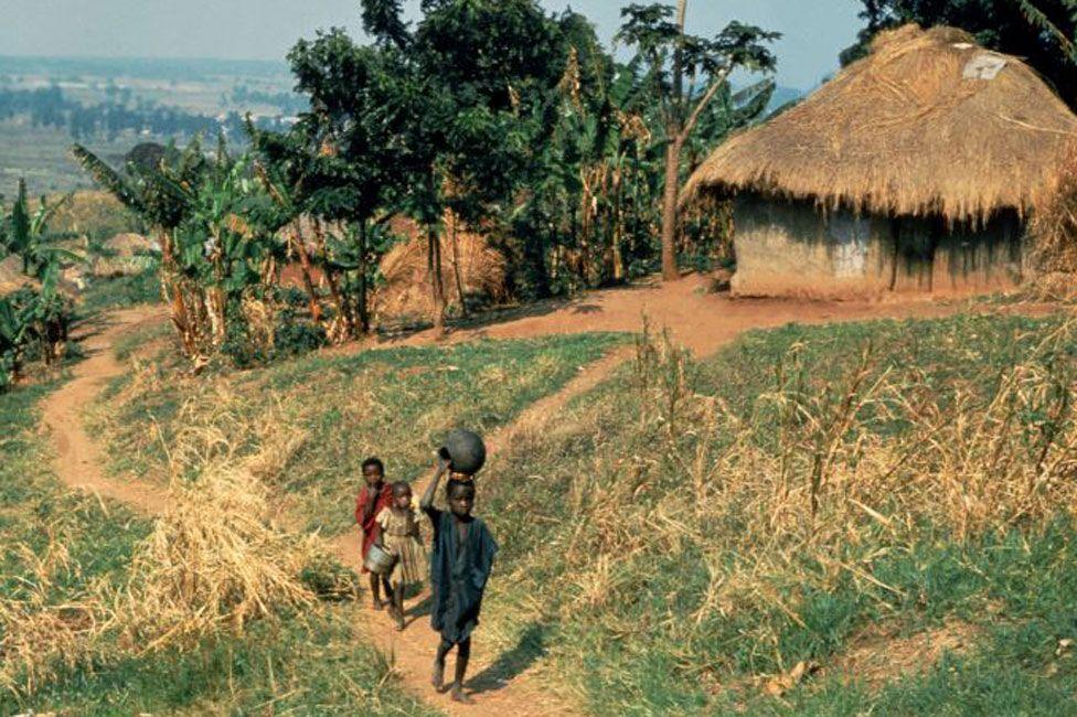 children walking on dirt path past thatched roof house