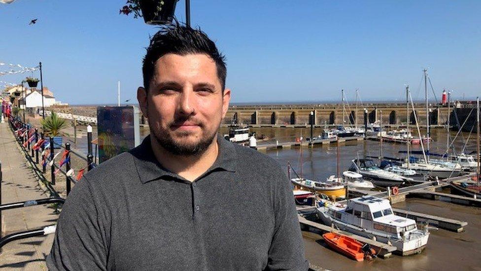 a white man with dark hair and jumper by a harbourside