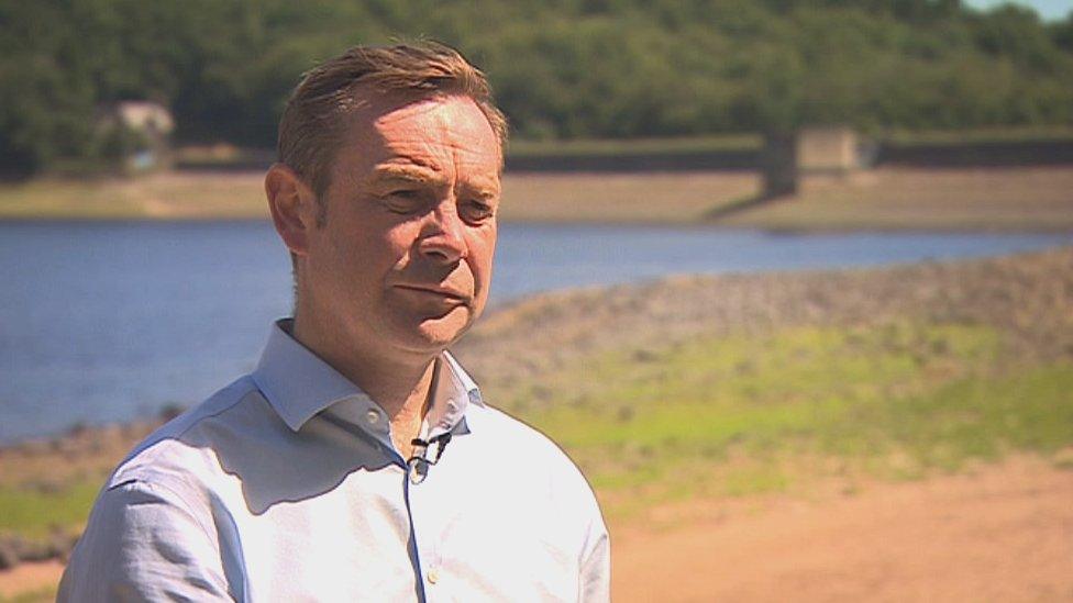 Image of a man stood with a reservoir behind him on a sunny day