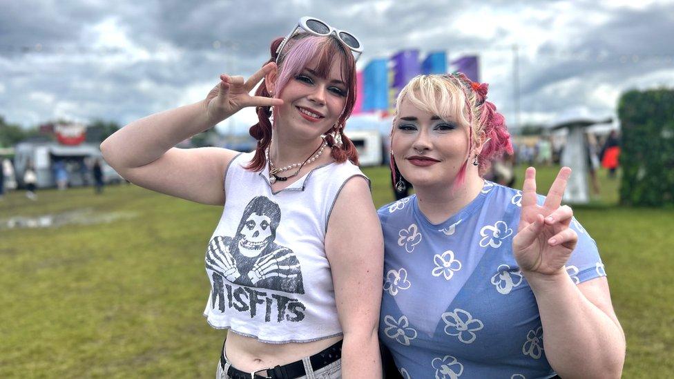 Yasi and Emily pose with peace signs at 90s Baby Festival, they're smiling. Yasi is wearing a white Misfits band t-shirt with the band's name written in a spooky font below a grim reaper character. Emily's wearing baby blue sheer top. They both have pink highlights in their hair. In the background we can make out a field and various food vans.