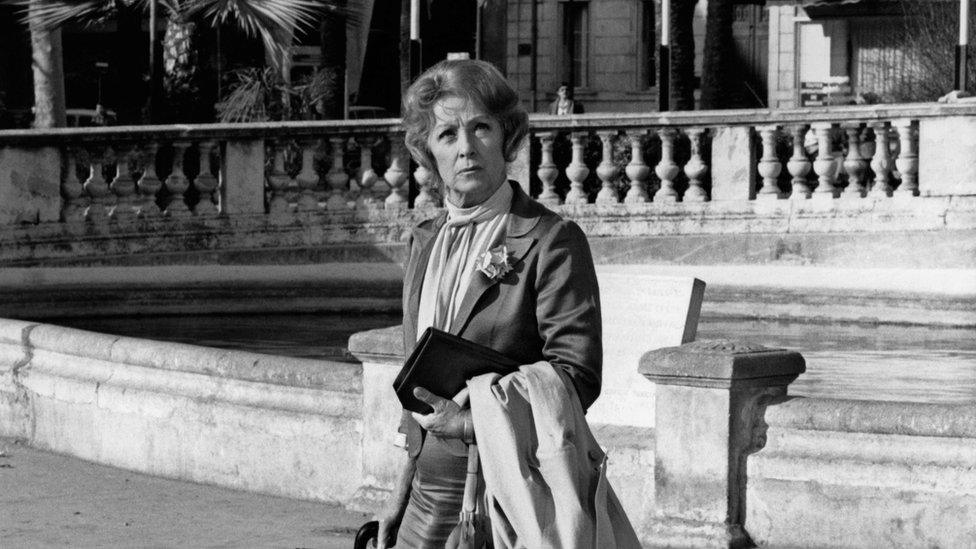 A woman in a smart blazer clutches her wallet with her coat on her arm