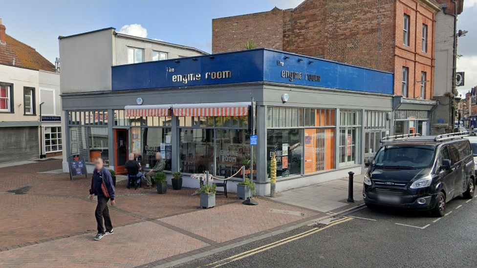 A single storey building with big window frontage on two sides, and blue backed signs above which say the engine room, some cafe tables and chairs outside the door