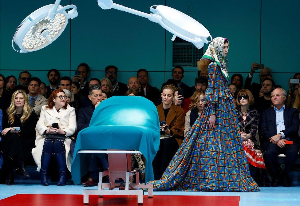 Female model in a brightly coloured dress and headscarf walks down the catwalk next to a medical bed as an audience looks on