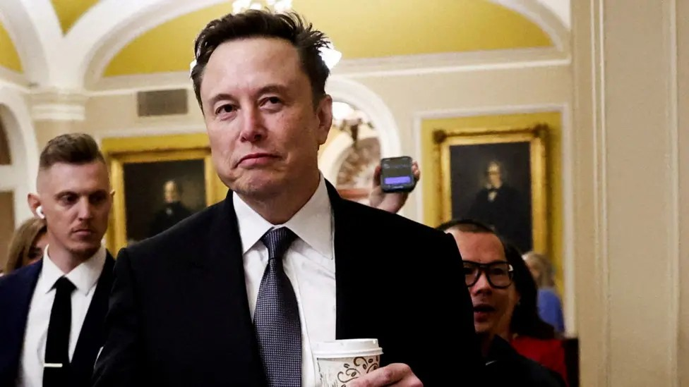 Elon Musk walks through Capitol Hill holding a takeaway coffee in his hand and looking away from camera, wearing a suit and tie and followed by security officers and journalists, in Washington DC on 5 December.