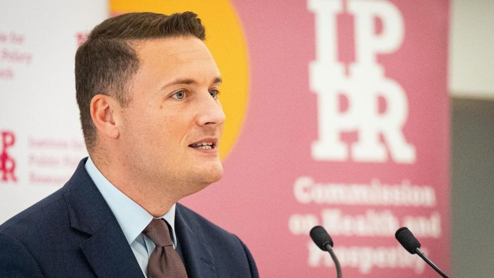 A side profile of Health Secretary Wes Streeting stood at a podium speaking into a microphone wearing a navy suit, blue shirt and brown tie. He is clean shaven with short, dark hair.