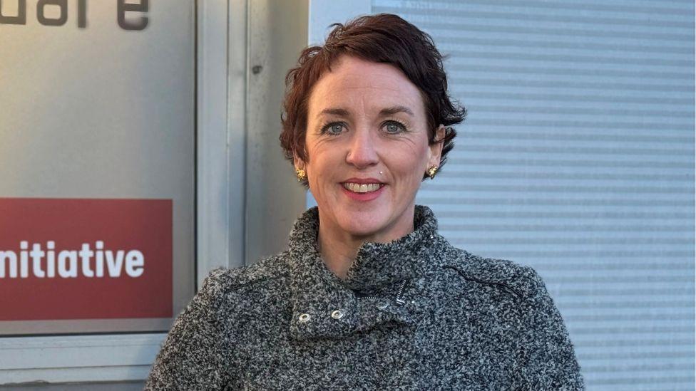 A woman, with dark hair and gold earrings, stands smiling at a camera in front of a metal sign. She is wearing a grey jumper 