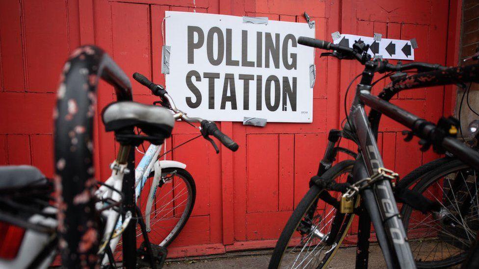 Two bikes are chained up outside a polling station in front of a bright red wall.