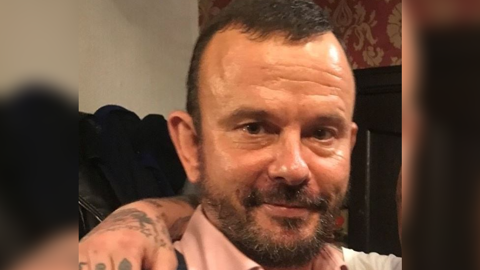 A head and shoulder photo of Eddie Roberts looking into the camera. He has a dark beard and short dark hair. He is wearing a pink shirt and has someone's arm over his shoulder. 