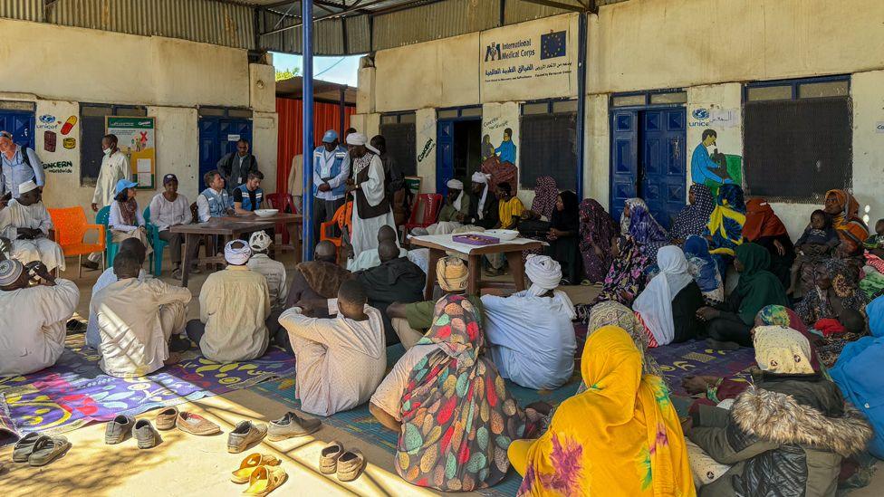 People with their backs to the camera sit on mats on the floor under a shelter listening to officials at the front sitting behind a table.