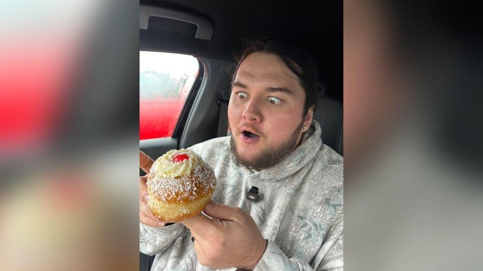 A man eating a doughnut with a cherry and cream on it. He has a beard and an earring in. He's wearing a white hoodie.