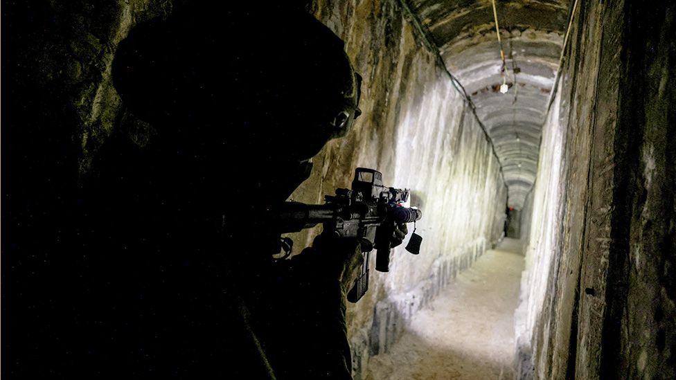 A soldier in dark tactical gear is holding a rifle with a mounted flashlight, illuminating a narrow, dimly lit tunnel with rough stone walls. The tunnel extends into the distance, curving slightly to the right.