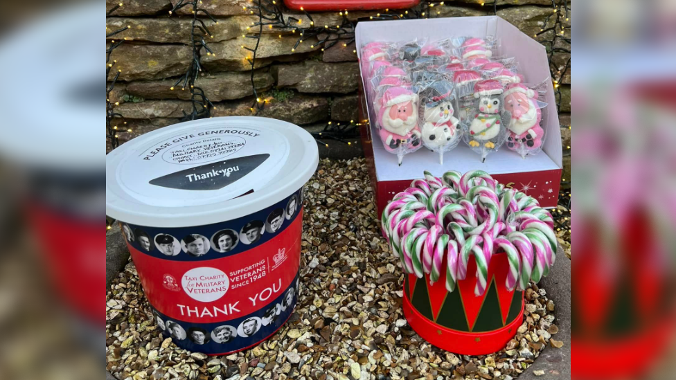 A donations box for Taxi Charity for Military Veterans featuring images of World War Two era servicemen and  women sits next two a box of candy canes and a box of lollipops in the shape of Father Christmas, snowmen and penguins. 