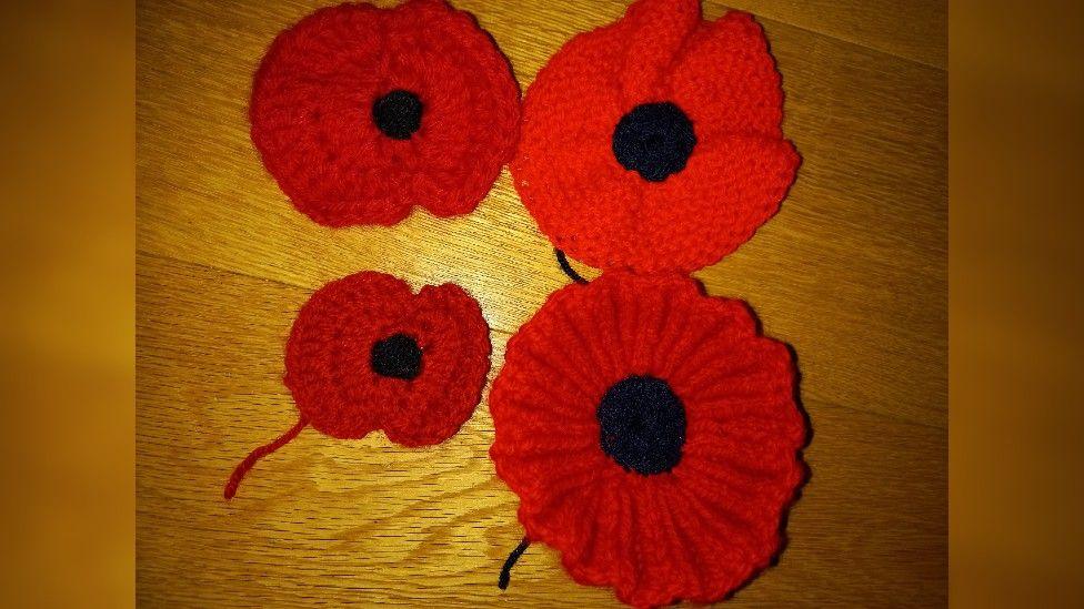 Four different crocheted poppies. One is circular with frills. One is circular without frills or undulation. The third is large and a similar shape to the poppies people can buy from the Legion, with indentation between the two large petals. The fourth is similar to the third, but much smaller, with a pronounced indentation. All have black middles, like the seeds on a poppy.