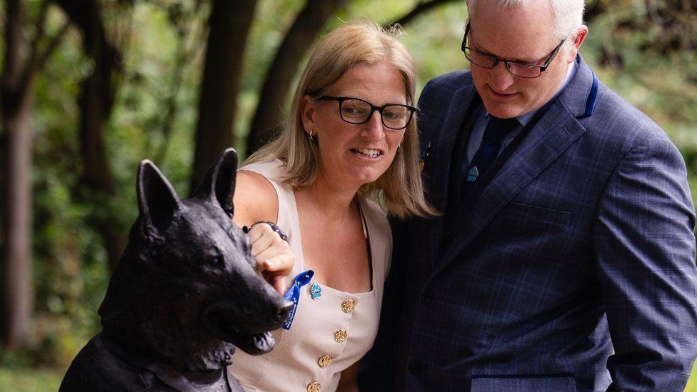 Two people looking at a statue of a German shepherd dog. On the left is the head of the dog, its head tilted to the left. In the middle is a woman with fair shoulder-length hair and wearing dark framed glasses and a white sleeveless dress pointing to the dog's nose. On the right is Dave Wardell, looking down at the statue, with white cropped hair, wearing dark framed glasses and a mid-blue suit, light blue shirt and dark blue tie. Behind are blurred out trees.