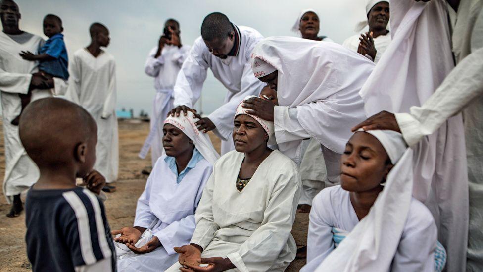 Three female apostolic church members sit with their legs in front of them and their palms upturned on their laps to receive blessings from fellow congregants who place hands on their heads. They are gathered outside in a rocky area in Harare and everyone is dressed in white expect for a child looking on and another child being held by a man in the background.