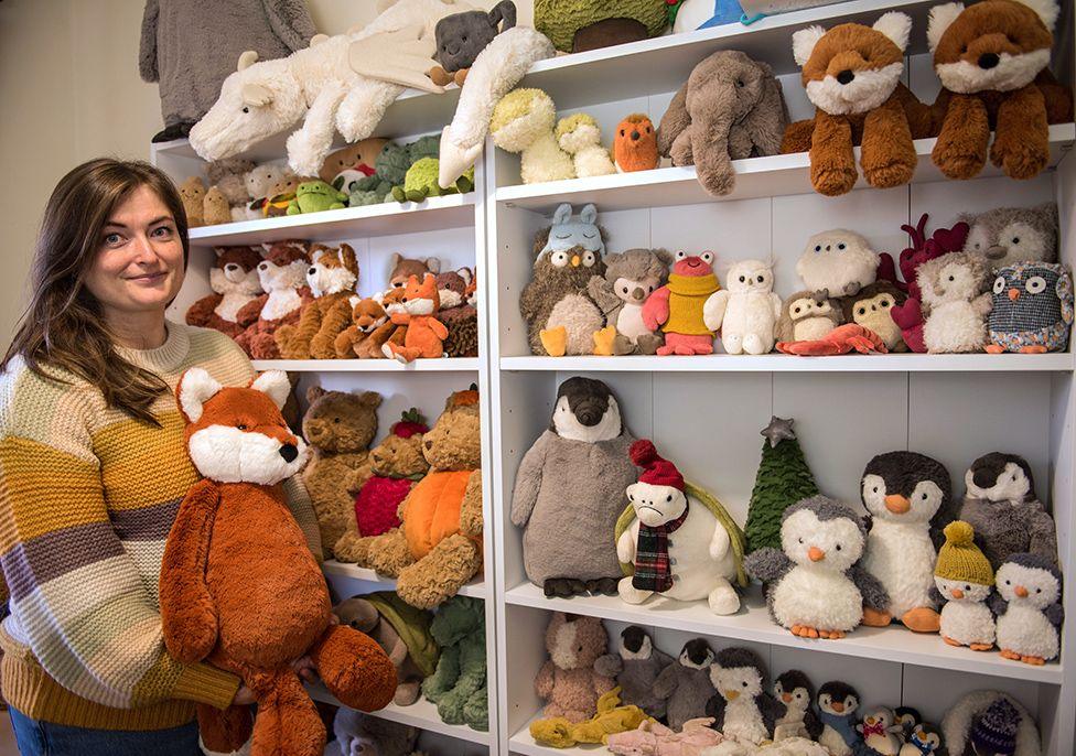 Amanda Hope stands holding a toy fox next to her collection of Jellycat toys stacked on shelves