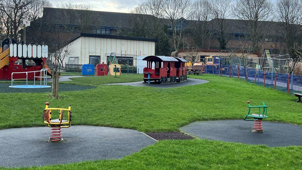 General view of the children's play area, a flat grassy area with a train, climbing frame and chairs on springs