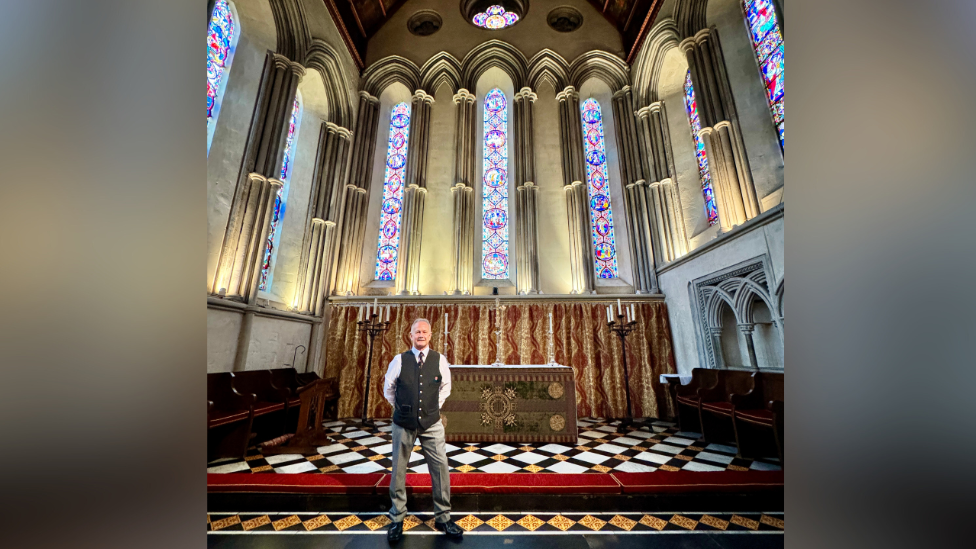 Dean Allen stood on impressive black and white checkered flooring inside a church. Mr Allen, who has his hands behind his back, is standing to face the camera while smiling. Behind him, above an alter, are three large arched stain glass windows. He is wearing his porters uniform. 