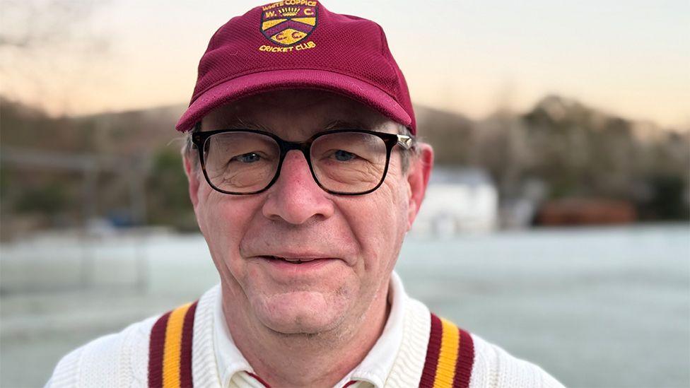 Head and shoulders image of club chairman, Paul Dobson standing on a frost covered cricket pitch. He is wearing glasses, is smiling and is wearing cricket whites and a burgundy cap