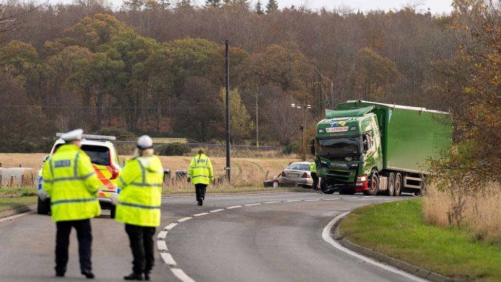 Police officers in yellow high-vis jackets at scene of crash, a green lorry and a grey car are in the distance on the bend of a rural road. There are fields and trees in the background.