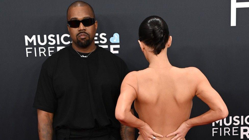 Kanye West wearing black opposite his wife wearing very little