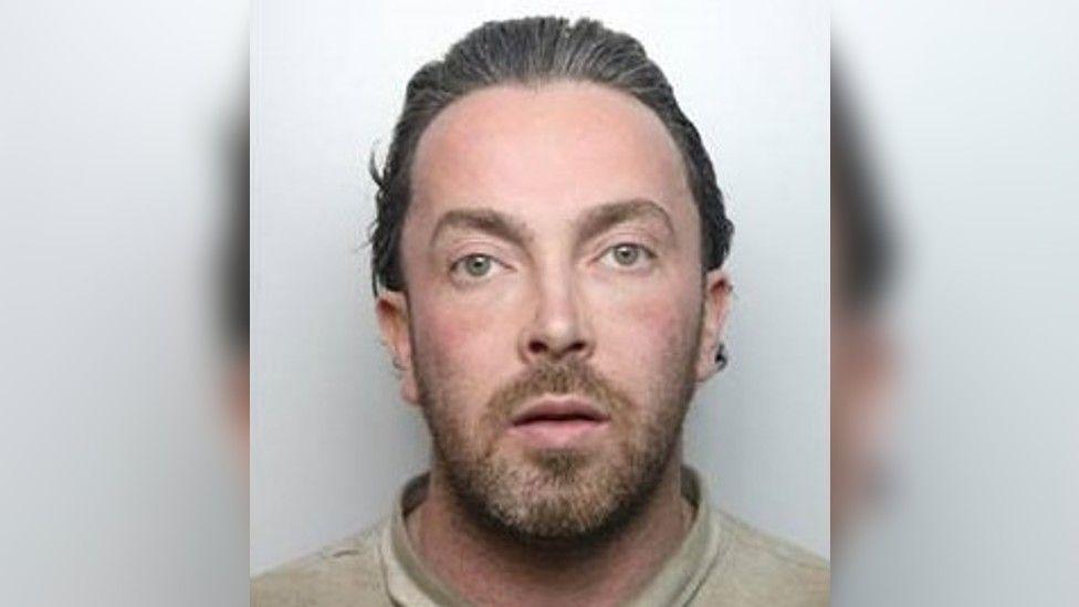 A police custody photo of a man with light brown hair and stubble, wearing a beige top.