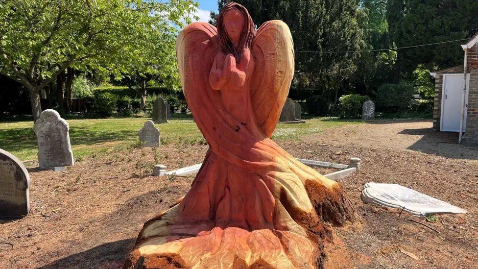 A carved wooden angel made from the stump of a tree. The angel has an orange coloured tan caused by preserving oil. Around the angel are various gravestones with more trees in the background.