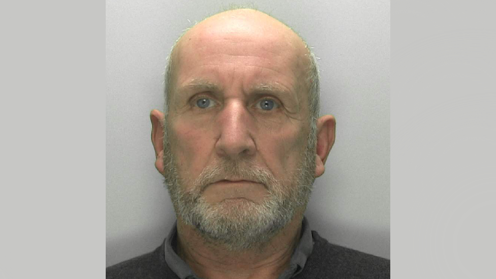 A police mugshot of Michael Florek. He has a bald head with grey hair on the sides, and a grey beard. He is wearing a dark collared shirt and standing against a grey background with a blank expression.