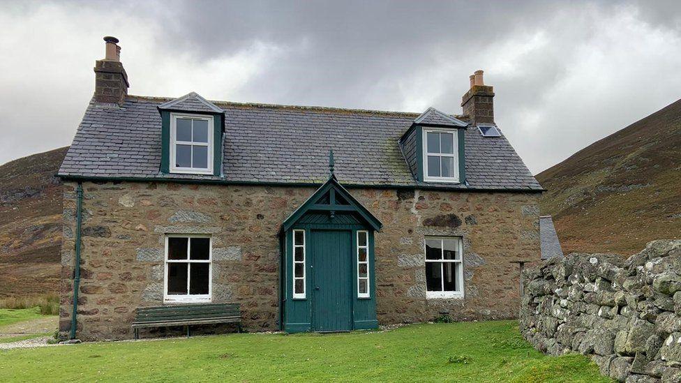 A stone cottage in the countryside with a green door and a wall to its right