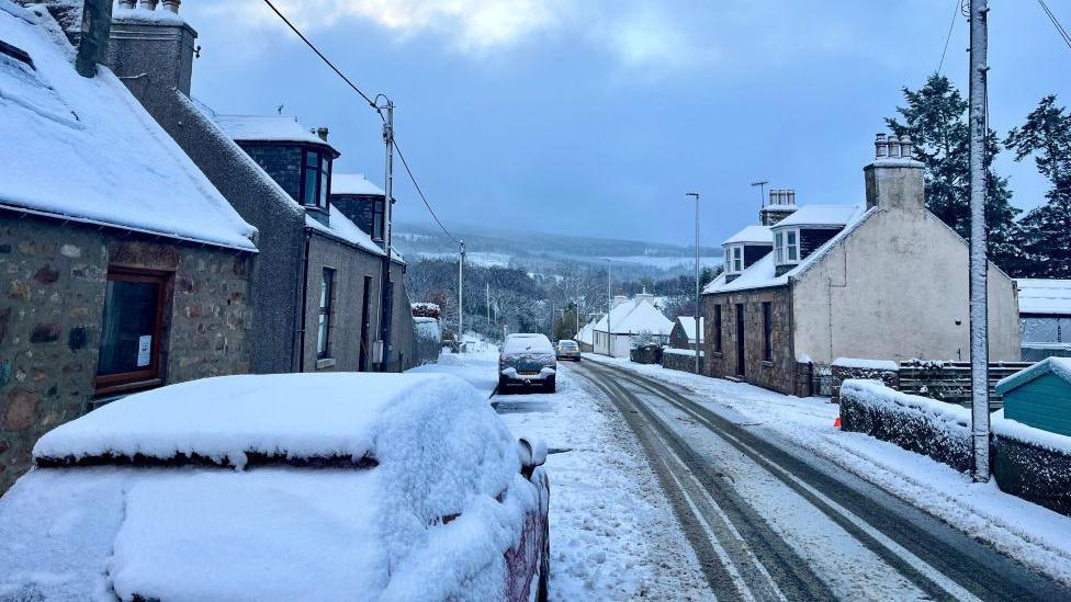 An image of a street and cars covered in snow in Auchleven in Aberdeenshire.