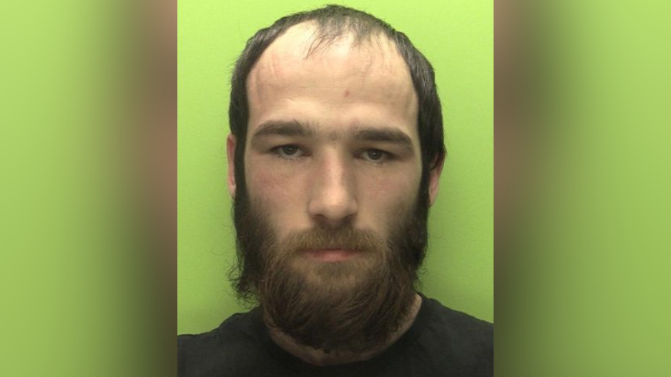 A mugshot of Jordan Wood, a white man with thinning dark brown hair and a beard, wearing a black t-shirt 