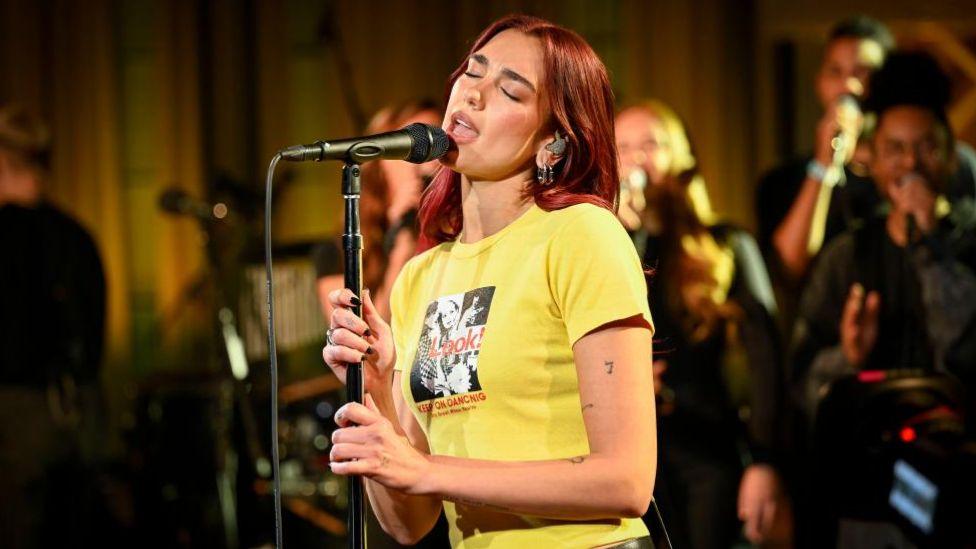 Dua Lipa stands with her eyes closed and sings into her microphone with musicians in the background. She is wearing a yellow T-shirt with a dance logo