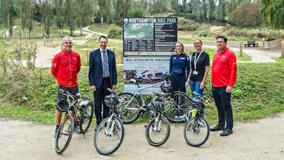 A line of three men and two women at a bike park with five bikes and helmets. They are standing in front of a map of the bike park and cycle trails are visible behind them.