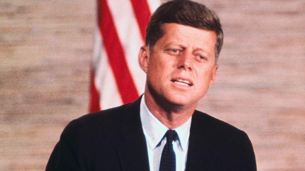 President John F. Kennedy, in a black suit, addresses at the podium in front of a US flag