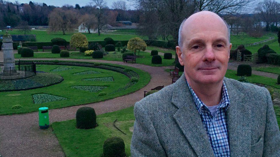 Oliver Wicksteed with short grey hair, wearing a grey jacket over a white and blue checked shirt.  He is standing in front of a maze-like garden with paths around small dome hedges and trees.