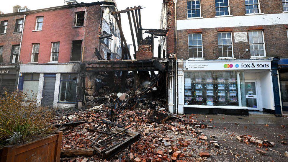 Building in centre of terrace with charred timbers sticking up on the first floor, bricks and burnt wood are flung across street.