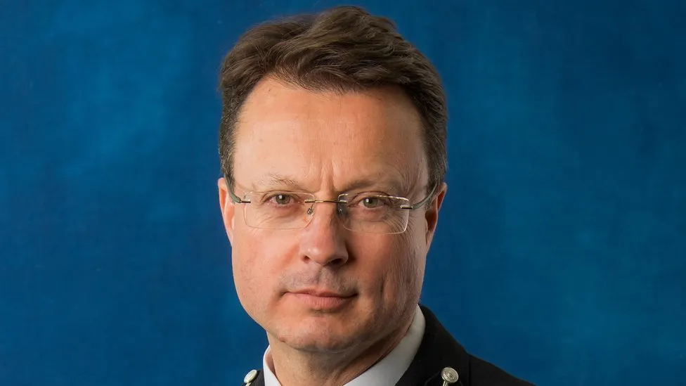 A close-up of Robin Smith looking at the camera wearing a police uniform. He has short brown hair and glasses