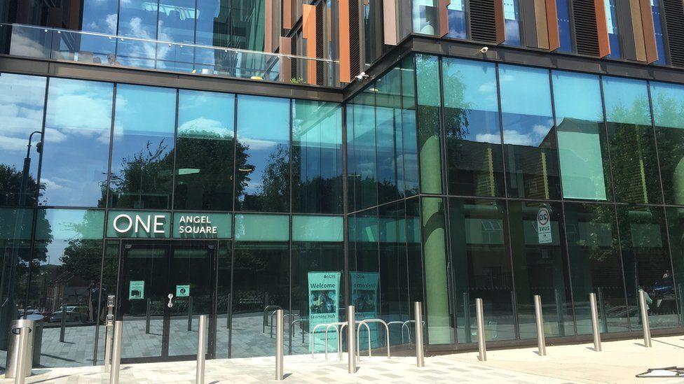 The headquarters of the Northamptonshire Children's Trust in Northampton. The entrance and lower part of the building is pictured with large glass panels. One Angel Square is written in large white letters by the entrance.