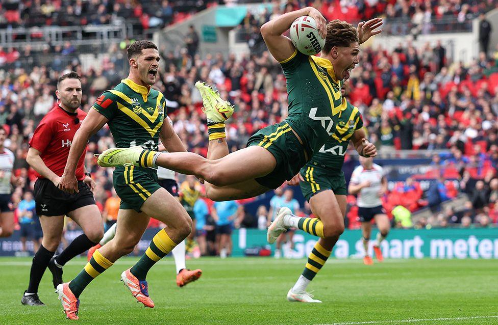 A rugby league player in a green and gold uniform leaps dramatically forward while holding the ball in one hand, scoring a try. Another teammate runs behind, the referee in  a red jersey is nearby. The background shows a packed stadium with spectators.